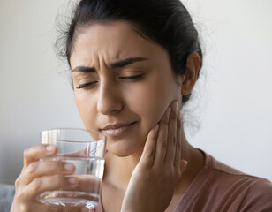 Young woman holds her jaw with a pained expression while bringing a glass of water to her lips, conveying toothache, dental discomfort and the search for relief at home.