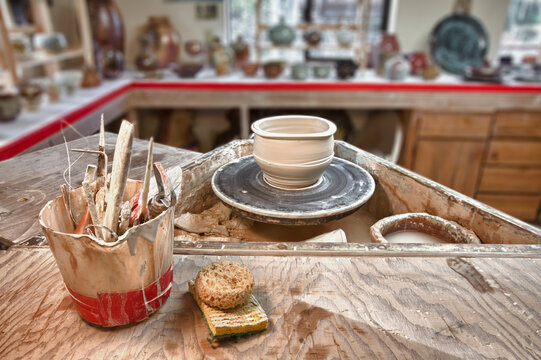 A potter's workshop, potters wheel, working tools and sponges on a board, displays of completed work in the background.