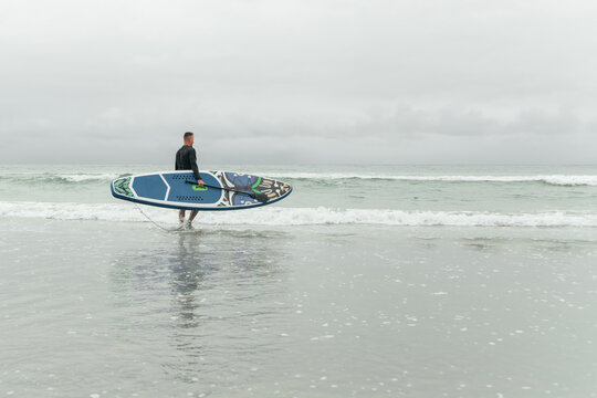 Surfer entering the ocean with paddleboard on overcast day.