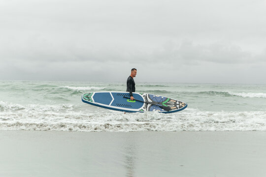 Smiling surfer entering the ocean with paddleboard on overcast day.