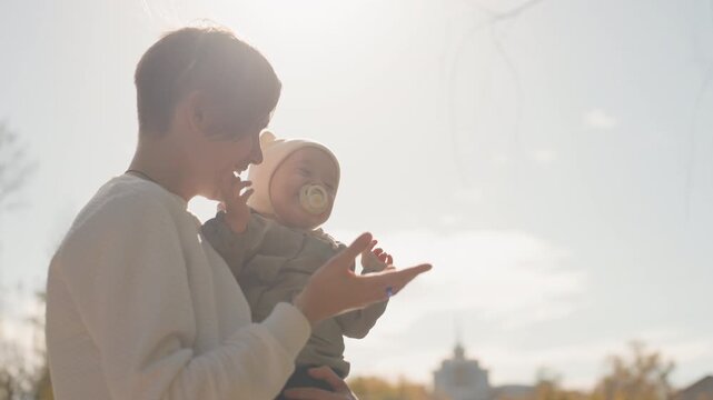 White father holding baby outdoors autumn. Soft backlight near green fence, knit beanie with ears, pacifier, gentle smile, close embrace, candid caregiving moment, parenting warmth and family