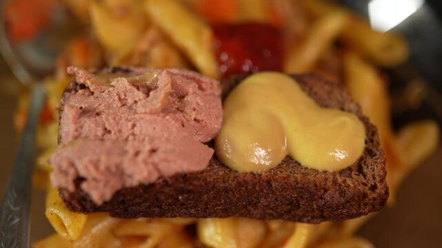 Close up shows rye bread topped with pate and glossy mustard over blurred penne pasta. 