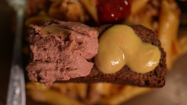 Close up shows rye bread topped with pate and glossy mustard over blurred penne pasta. 