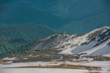 Sunlit mountain landscape with small alpine lake among sharp rocks and snowfields with view to forest hills. Rocky outcrops among snows and thawed ground during thaw. Sunlight and shadows of clouds. © Daniil