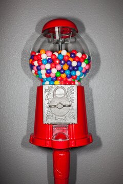 Close-up of a red gumball machine filled with vibrant multicolored gumballs against a gray textured wall. Fun and nostalgic candy concept.
