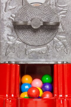 Colorful gumballs visible inside the dispensing slot of a red vintage gumball machine with textured metal turn handle.