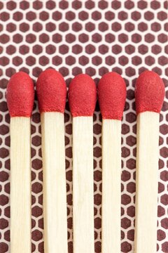 Close-up macro image of five unlit wooden matchsticks with red heads arranged side by side on a brown matchbox striking surface, the hexagonal pattern of the striker panel. 
