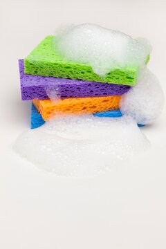 Brightly colored household sponges stacked and covered in foamy soap bubbles against a clean white background. 