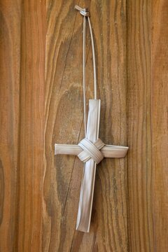A simple handmade cross crafted from dried palm fronds hangs against a rustic wooden background. This traditional cross, often associated with Palm Sunday