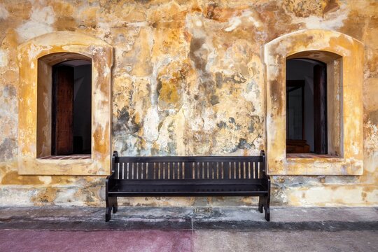 A simple wooden bench sits between two arched windows against a textured, weathered wall inside Castillo San Cristobal in Old San Juan