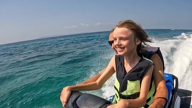 A child in a life jacket rides a jet ski with an adult in the open sea &ndash; a sunny day, water splashes and waves in the background.
