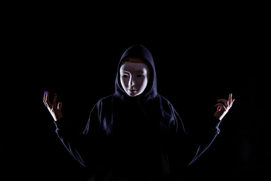 Person wearing a white expressionless mask and dark hoodie standing against a black background with raised hands in a dramatic gesture. Strong contrast lighting create a mysterious atmosphere