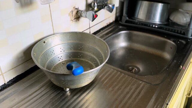 Old metal colander holding a blue cleaning sponge placed on a stainless steel drainboard next to a kitchen sink with wall faucet and tiled background in natural indoor light.