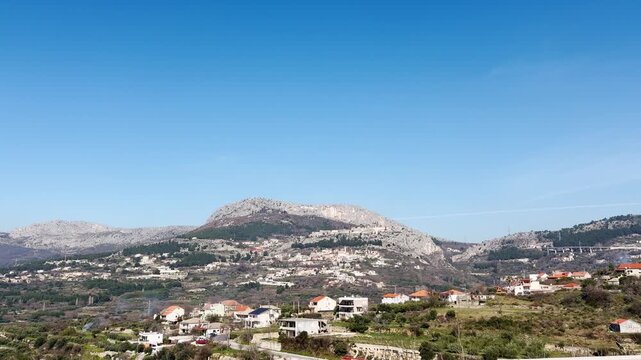 Side view from a moving car passing Klis near Split, showing hillside homes, the Klis Fortress on a ridge, and clear blue sky during a Croatia road trip.