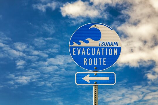 Blue and white tsunami evacuation route sign with arrow, photographed against a bright sky with scattered clouds in Bandon, Oregon.