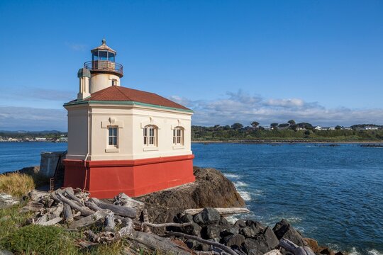 Historic Coquille River Lighthouse in Bandon, Oregon, stands on rocky shore with driftwood in foreground and blue coastal waters under a bright, sunny sky.
