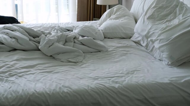 Unmade bed with rumpled white sheets and pillows, showing gradual disarray in bedding and a dark garment draped on the edge in a well-lit bedroom setting