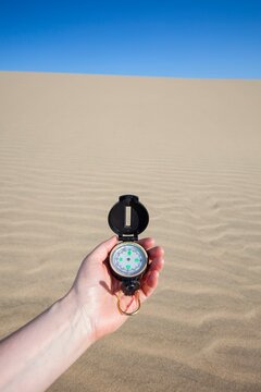 A persons hand holding a navigation compass against the backdrop of a sandy dune under a vivid blue sky, symbolizing exploration, direction, and outdoor adventure.