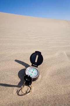 A navigation compass rests on a sandy dune beneath a vibrant blue sky, representing themes of travel, exploration, navigation, and adventure in a desert or coastal environment.