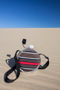 A striped fabric-covered canteen with a shoulder strap rests on the rippled surface of a sandy dune 