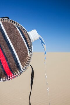 Close-up of a striped fabric-covered canteen tilted to pour out a stream of water, with the backdrop of a sandy dune and blue sky, representing survival, scarcity, and hydration in arid environments.