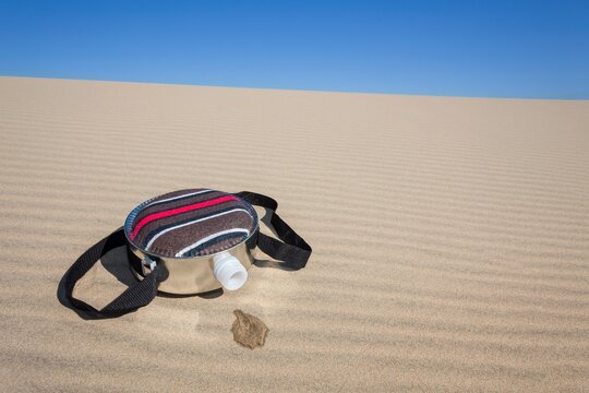 A fabric-covered canteen rests on rippled sand dunes with a small puddle of spilled water soaking into the dry sand, symbolizing survival and scarcity in desert environments.