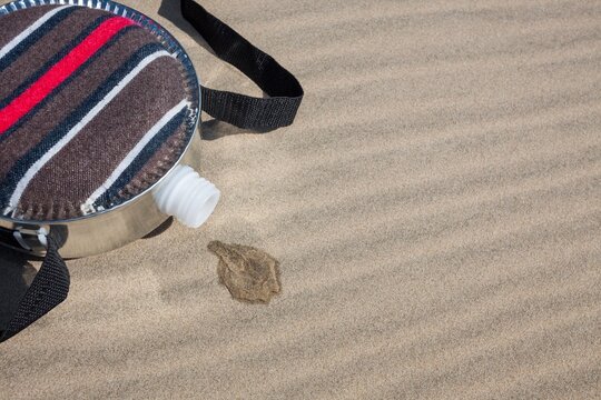 A fabric-covered canteen rests on rippled sand dunes with a small puddle of spilled water soaking into the dry sand, symbolizing survival and scarcity in desert environments.