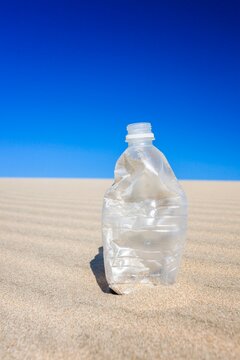 Crushed, empty plastic water bottle standing on rippled desert sand under a hot blue sky, evoking thirst, heat, and survival.