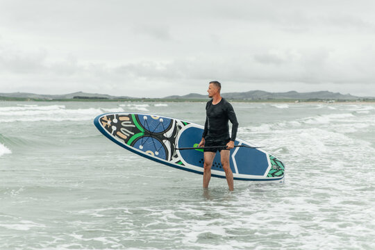 Man standing in water holding paddleboard and paddle.