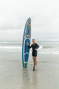 Man standing in water holding paddleboard and paddle.