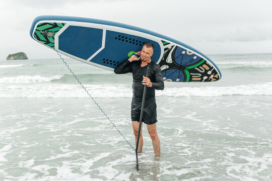 Smiling man standing in waves with paddleboard and paddle.