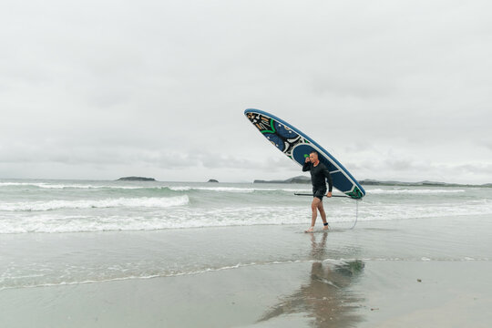Man walking on beach carrying a paddleboard on shoulder.