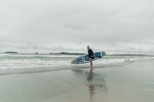 Man walks along shore with paddleboard on cloudy day.