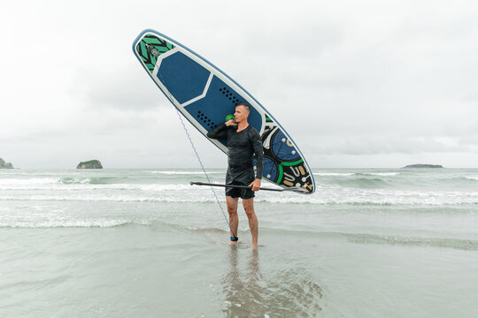 Surfer holding paddleboard on shoulder, smiling at beach.