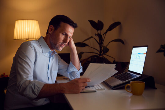 Tired businessman working overtime at home desk with documents and laptop