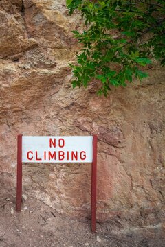 Warning sign reading "No Climbing" in red letters, mounted on wooden posts, placed against a rugged rock wall with green leaves overhead, Colorado Springs, Colorado