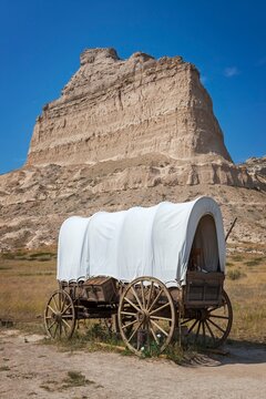 A classic covered wagon known as a prairie schooner, on open prairie below the towering sandstone bluffs of Scotts Bluff National Monument in western Nebraska on the Oregon trail.