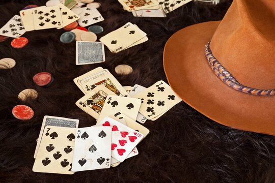 Weathered playing cards and clay chips scattered on dark animal hide beside a cowboy hat, evoking a saloon poker game in an Old West town, gambling.