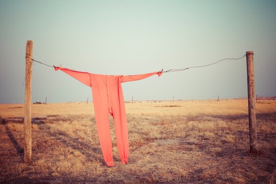 Bright red long johns hang from a wire clothesline between fence posts on a wide South Dakota prairie, with open sky and dry grass creating a simple rustic scene.