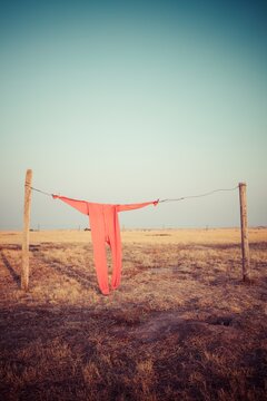 Bright red long johns hang from a wire clothesline between fence posts on a wide South Dakota prairie, with open sky and dry grass creating a simple rustic scene.