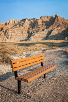 An empty bench faces the layered buttes of Badlands National Park, South Dakota, at golden hour. Prairie grass and a dusty trail lead toward dramatic eroded cliffs and a calm open sky.