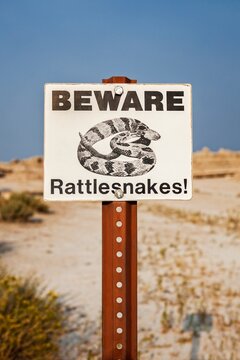 Close view of a Beware Rattlesnakes sign against blue sky and dry prairie in Badlands National Park, South Dakota, highlighting safety messaging and wildlife hazard awareness.
