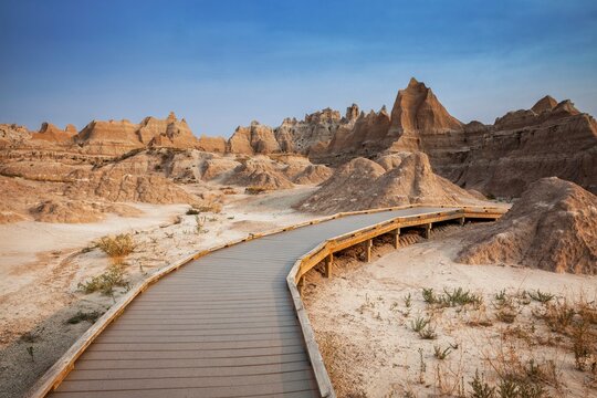 A curving boardwalk leads into eroded buttes and pinnacles under a clear blue sky in Badlands National Park, South Dakota, inviting exploration of a rugged desert landscape.