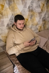 Obraz premium A man wearing a beige hoodie and black pants relaxing on a couch while using a mobile device