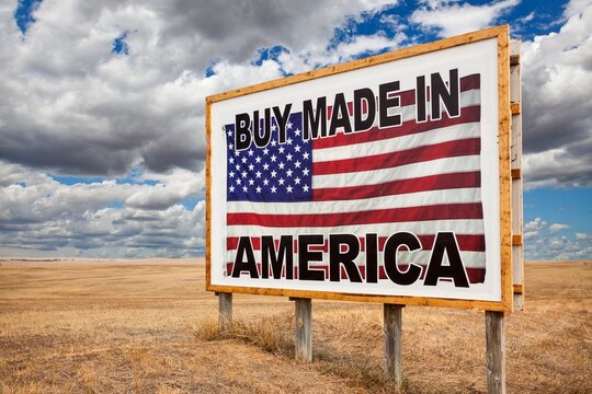 A large roadside billboard with an American flag reading Buy Made in America stands on a dry prairie under dramatic clouds in South Dakota; patriotic rural scene with open sky.