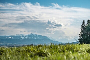 Obraz premium View from Mariaberg above Kempten toward the Allgäu Alps under mixed clouds, green meadow in the foreground and partially snow-covered alpine peaks in the distance.