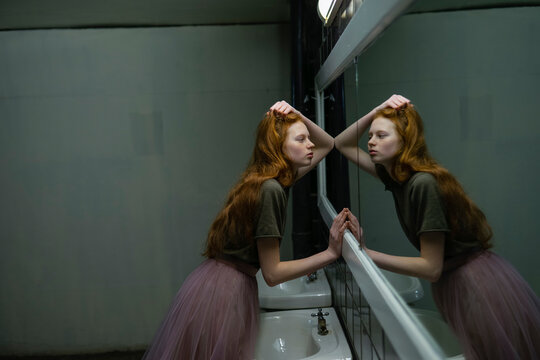 Red-haired woman with freckles leans on bathroom sink in front of mirror