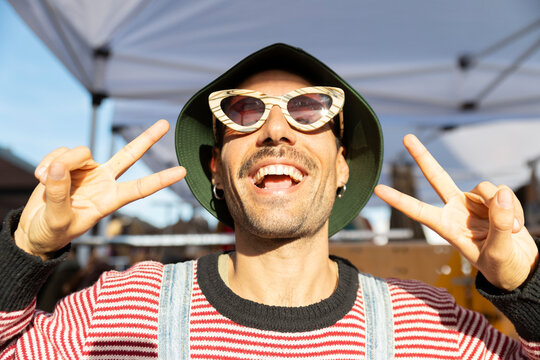 Playful man in sunglasses and hat making peace sign outdoors
