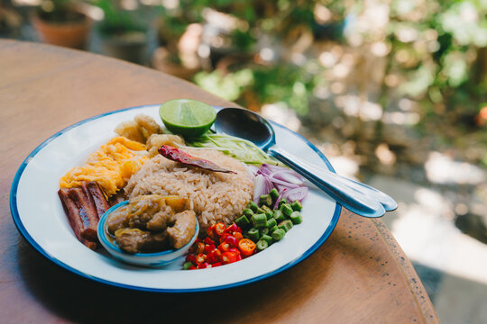 Kao kluk kapi Thai food. Rice dish with chili, lime, egg, and vegetables served on white plate with spoon and fork on wooden table in natural light
