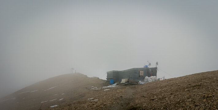Mountain refuge hut near summit cross on Punta Penia in foggy conditions with rocky terrain and snow patches in Dolomites Italy